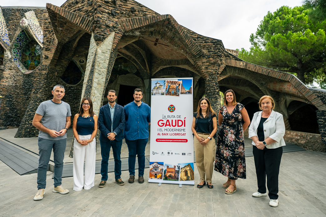 Els representants municipals i comarcals posen davant la cripta de Gaudí amb el cartell de la Ruta del Modernisme de la comarca